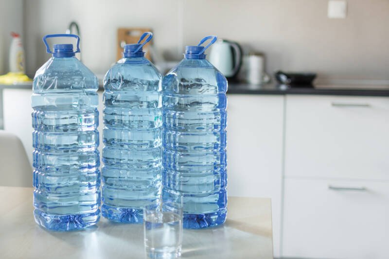 Una gran botella de plástico con agua en la mesa sobre un brillante fondo de cocina.  Botella de agua transparente transparente en una tapa de color azul y cierre de mango